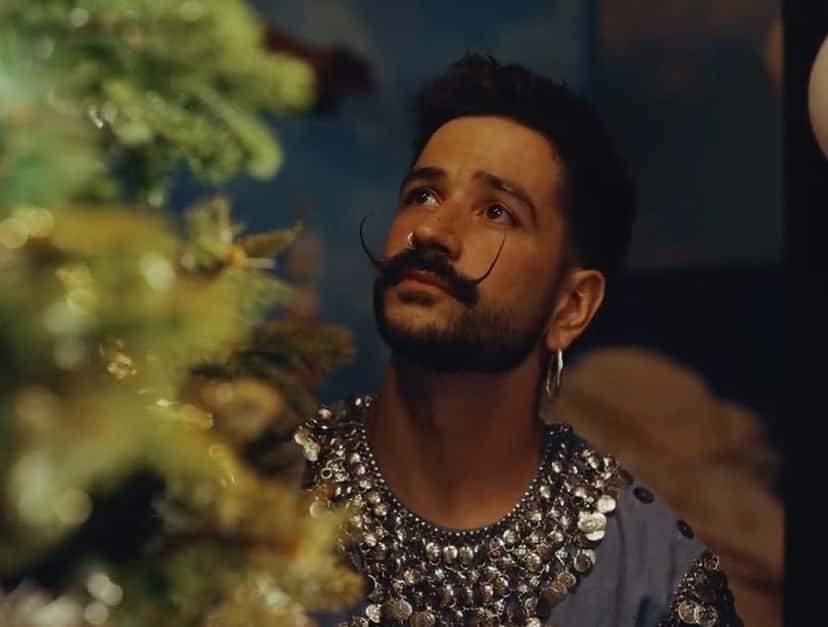 Camilo en el video oficial de «El Árbol»: de bigote largo y camisa con apliques plateados, mira hacia arriba junto a un árbol de Navidad (fotograma del video oficial)