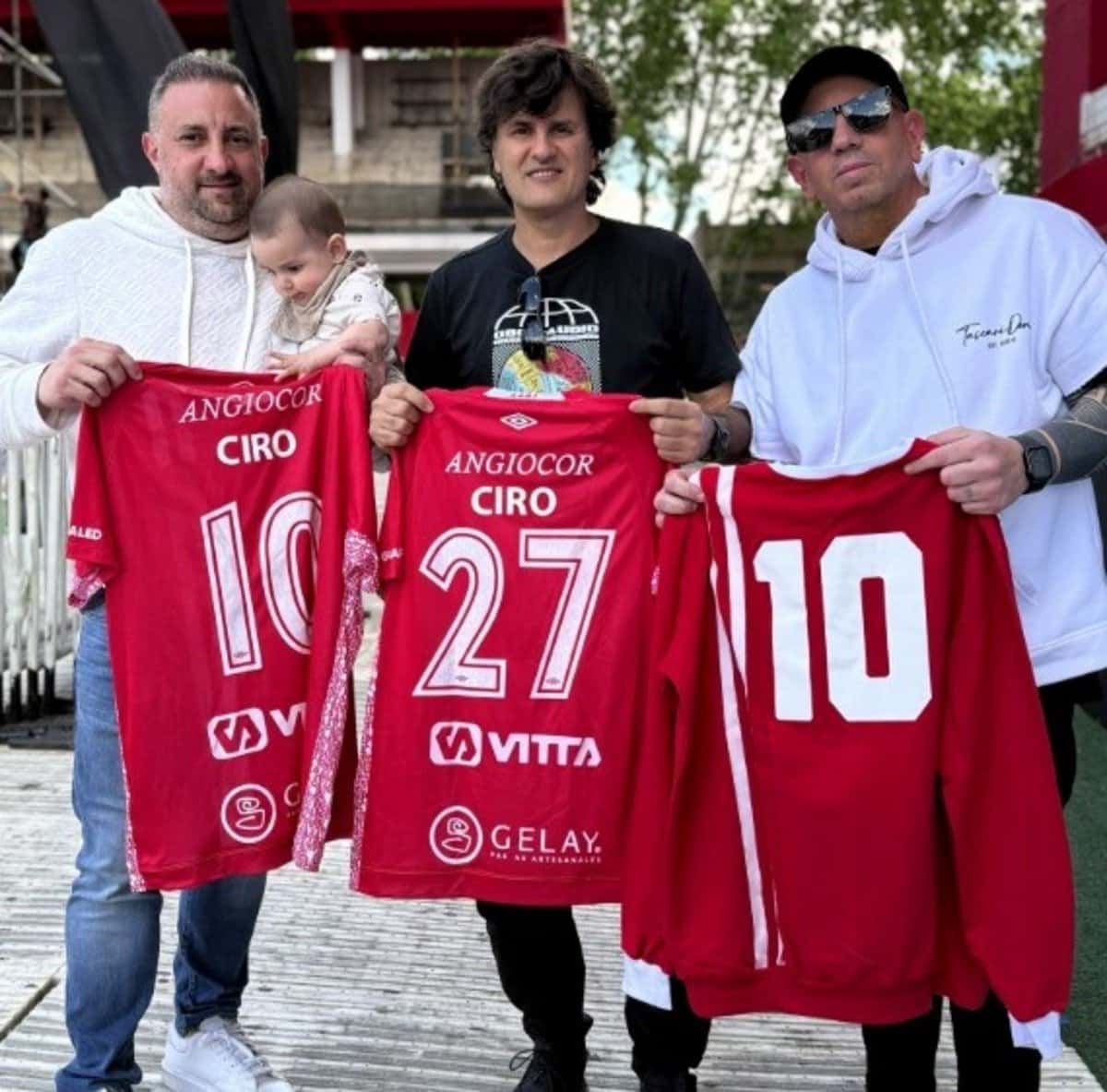 Andrés Ciro Martínez, en el centro, muestra una camiseta roja de Argentinos Juniors con su nombre y el número 27, junto a otras dos personas con camisetas del club, en la previa del recital en el estadio Diego Armando Maradona