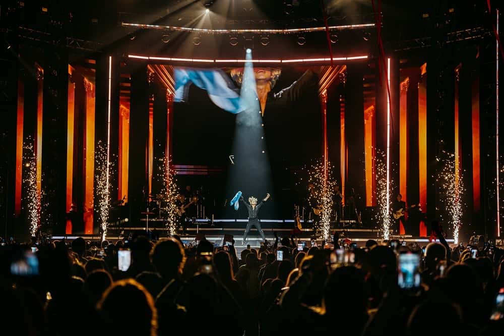 Foto de prensa oficial del show de David Bisbal en el Movistar Arena: el cantante, solo en el centro del escenario bajo un haz de luz, levanta una bandera argentina mientras otra flamea en la pantalla, con columnas iluminadas en naranja, chispas de fuegos fríos a los costados y el público con los celulares en alto en primer plano