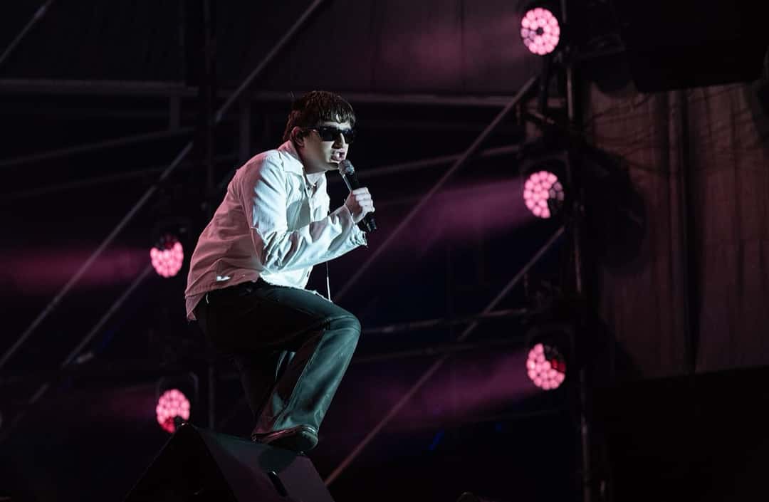 Dillom canta en cuclillas sobre un monitor del escenario, con camisa blanca, pantalón oscuro y anteojos de sol, bajo luces rosas, en el Cosquín Rock 2025 (foto de archivo). Foto: Juan Manuel Ramos, CC BY-SA 4.0, vía Wikimedia Commons