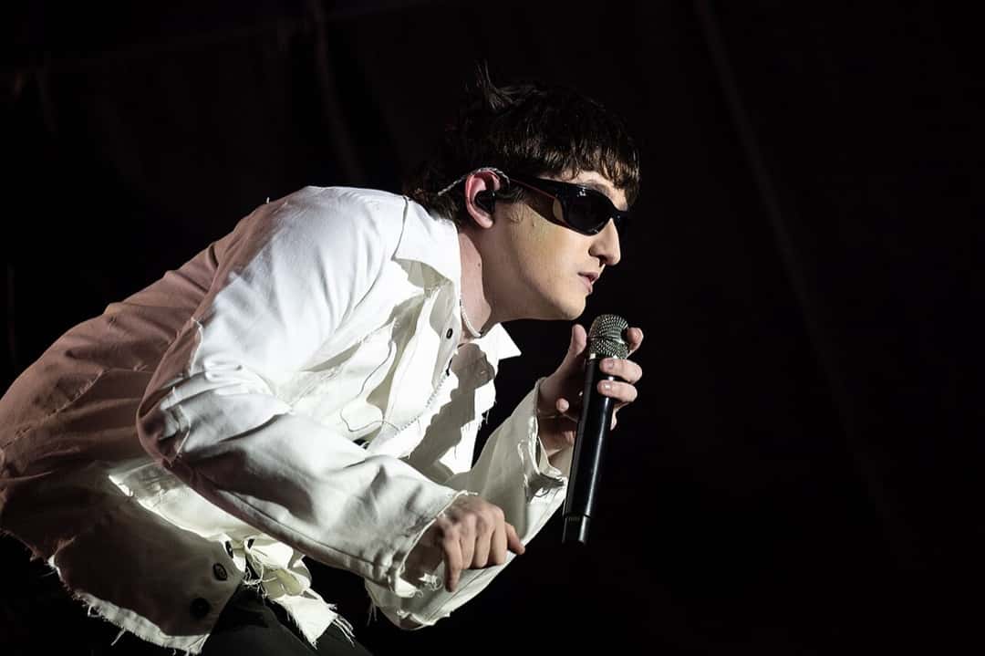 Dillom de perfil, con camisa blanca desflecada y anteojos de sol, canta inclinado hacia adelante con el micrófono en la mano, sobre fondo negro, en el Cosquín Rock 2025 (foto de archivo). Foto: Santiago Mazzarovich, CC BY-SA 4.0, vía Wikimedia Commons