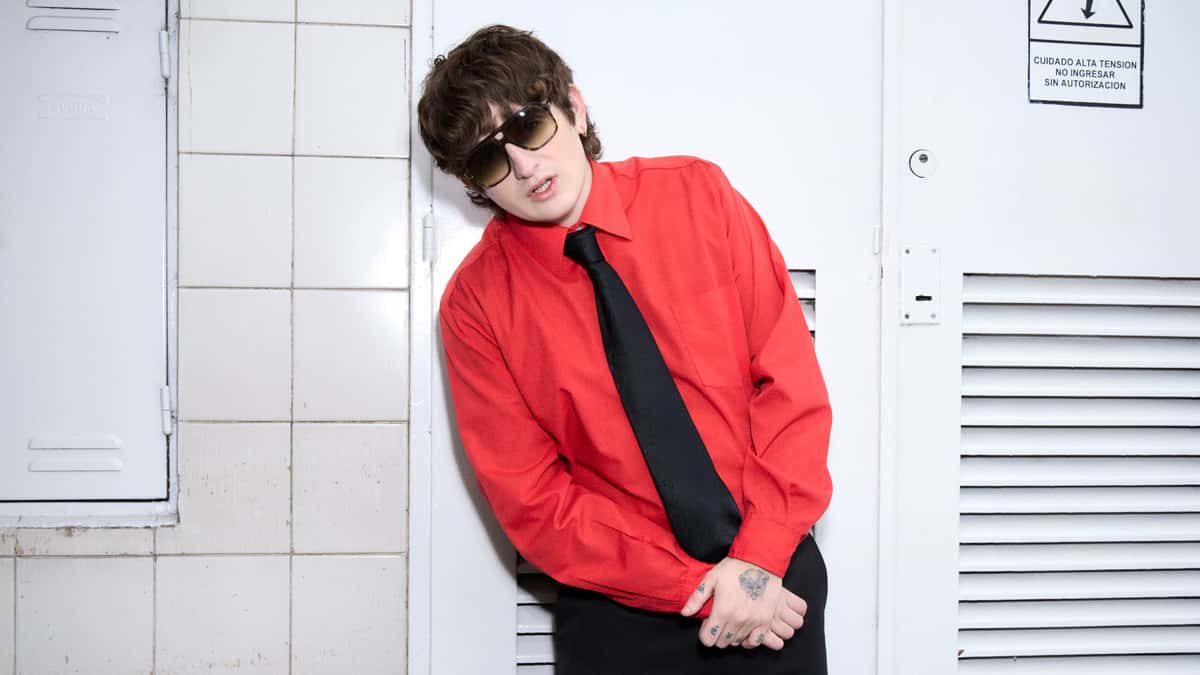 Dillom con camisa roja, corbata negra y anteojos de sol, apoyado contra una puerta metálica blanca, en la foto de prensa oficial de la etapa de «La Nueva Violencia»