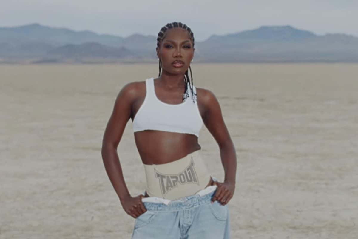 Imagen del video de «CATWOMAN»: Doechii, con trenzas, top deportivo blanco, faja con la palabra TAPOUT y jean ancho celeste, parada con las manos en los bolsillos en un desierto llano con montañas de fondo. Imagen: Doechii / YouTube