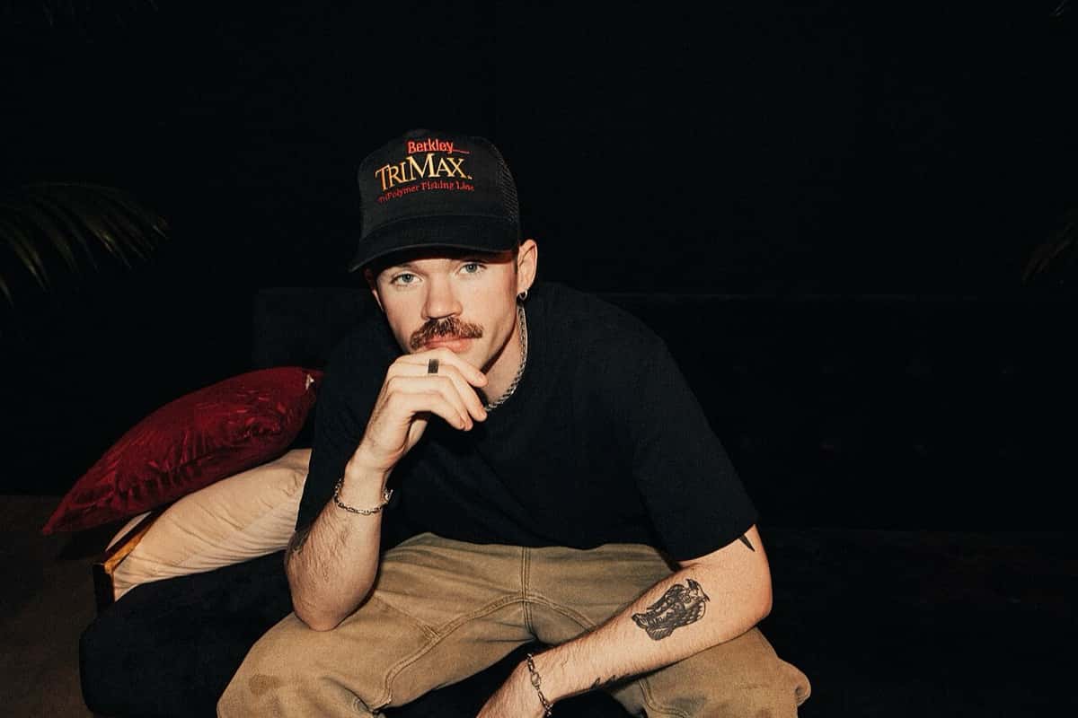 Foto de archivo de Dom Dolla sentado en un sillón oscuro, con gorra negra, remera negra y pantalón beige, apoyando el mentón en la mano