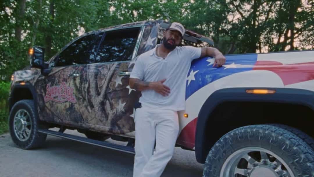 Drake con gorra y ropa blanca, apoyado en una camioneta pintada con la bandera de Estados Unidos, en una imagen de video