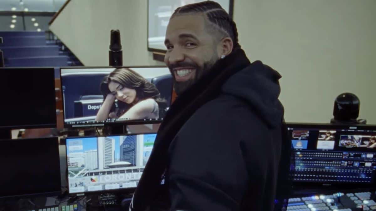 Drake sonriendo, con buzo negro con capucha, frente a una consola de transmisión con varios monitores encendidos