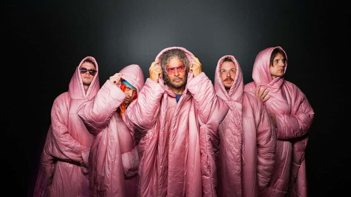 Los cinco integrantes de The Flaming Lips con capas acolchadas rosas con capucha, frente a un fondo negro; Wayne Coyne, al centro, con anteojos rosas