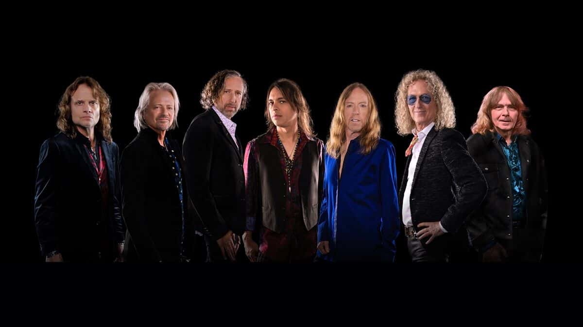Foto de prensa de Foreigner: siete músicos de pelo largo, con camperas y camisas oscuras, posan en fila sobre un fondo negro