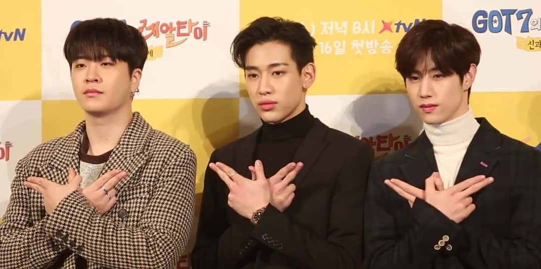 Tres integrantes de GOT7 posan con los brazos cruzados sobre el pecho frente a un fondo amarillo, en la presentación de un programa de televisión (foto de archivo, enero de 2019). Foto: K-POPIT 케이팝잇, CC BY 3.0, vía Wikimedia Commons