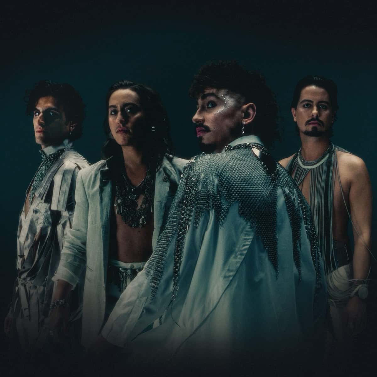 Los cuatro integrantes de Greta Van Fleet posando sobre fondo verde azulado oscuro, con ropa clara, cadenas y brillos en la cara