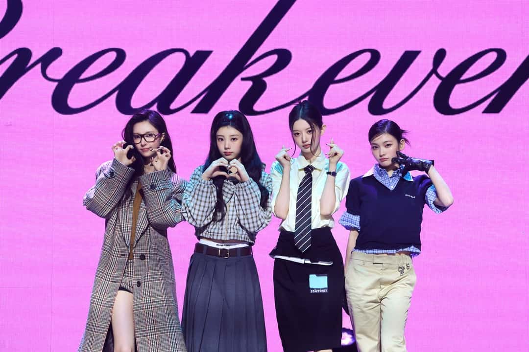 Las cuatro integrantes de ILLIT posan juntas frente a una pantalla rosa con el nombre «Break Even», haciendo corazones con las manos, en la presentación del disco en Seúl. Foto: Belift Lab