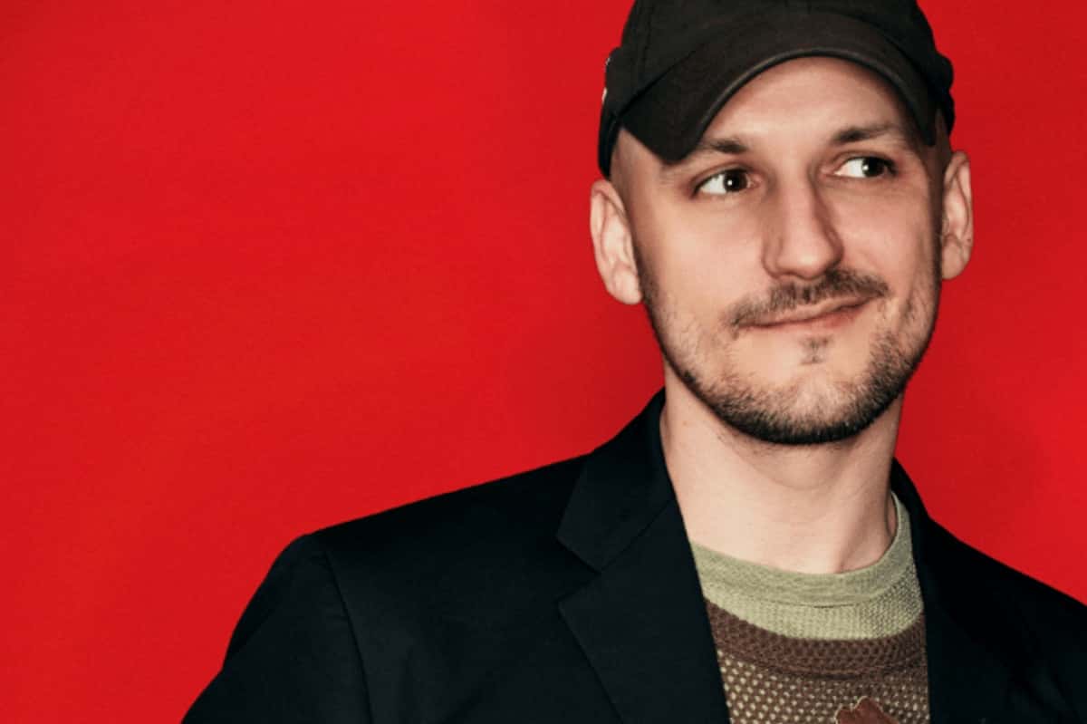 Retrato de prensa de Jacques Greene con gorra negra, saco oscuro y remera verde, mirando hacia un costado y sonriendo apenas, frente a un fondo rojo liso