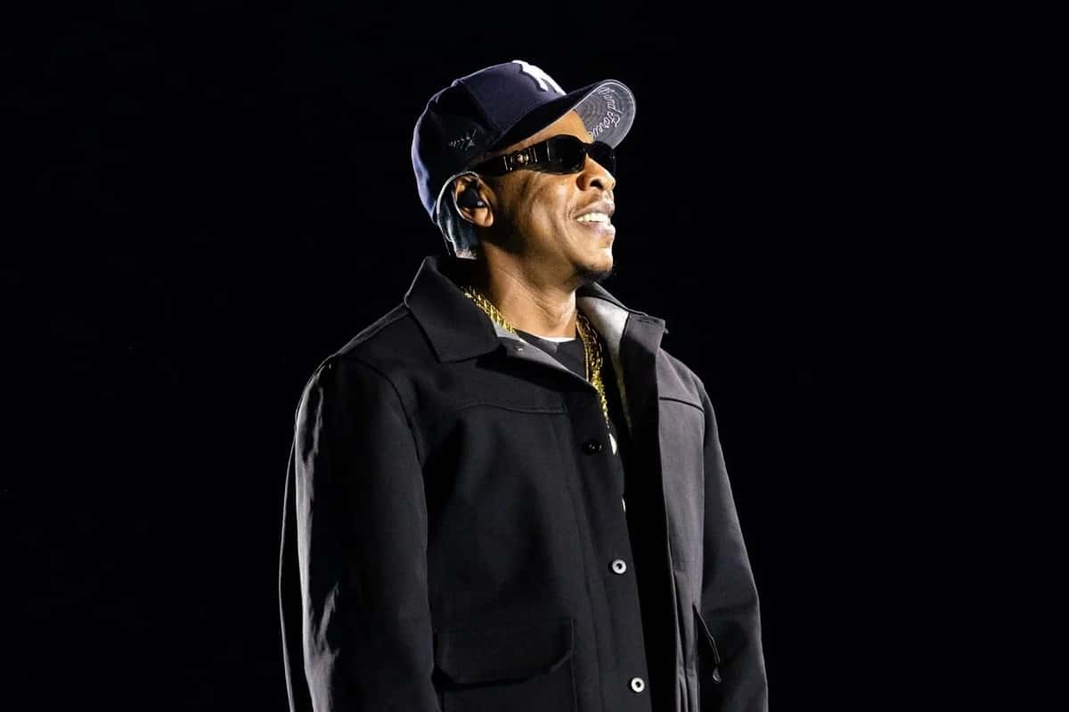 Jay-Z sonríe en el escenario del SoFi Stadium, con gorra azul de los Yankees, anteojos negros, campera oscura y cadena dorada, sobre fondo negro