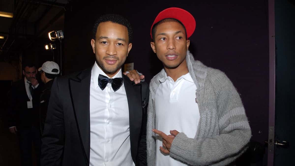 John Legend con esmoquin negro, moño y camisa blanca, posando junto a Pharrell Williams, que lleva gorra roja, chomba blanca y cárdigan gris tejido, en un backstage con fondo oscuro