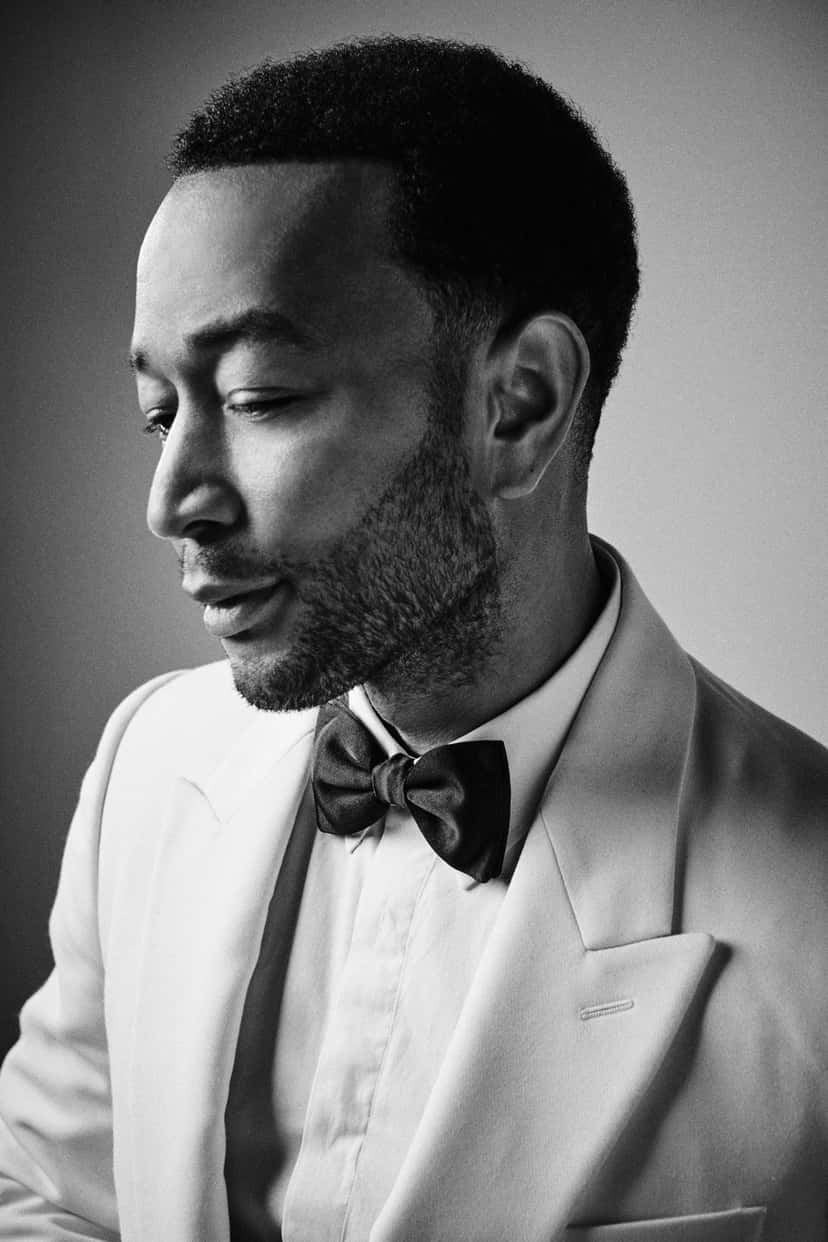 Retrato en blanco y negro de John Legend de perfil, mirando hacia abajo, con saco claro, camisa blanca y moño negro, sobre un fondo gris liso