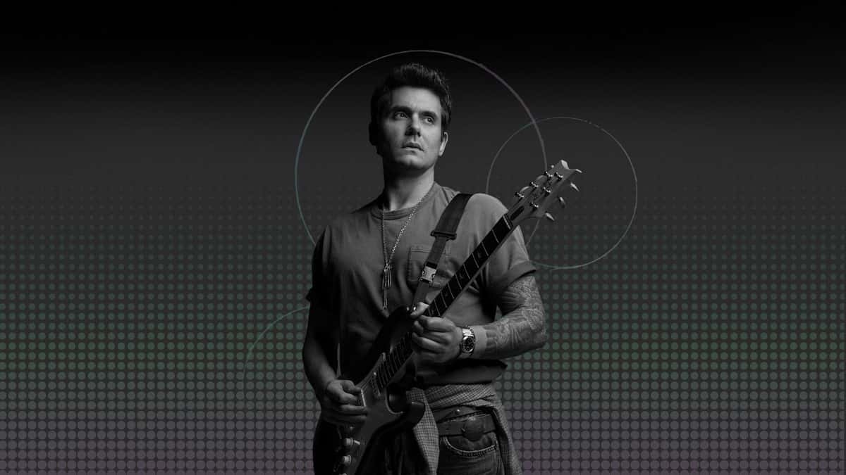 Foto oficial de la residencia de John Mayer en la Sphere: el músico, en blanco y negro, con remera, collar largo y una guitarra eléctrica, mirando hacia un costado sobre un fondo oscuro de puntos y círculos