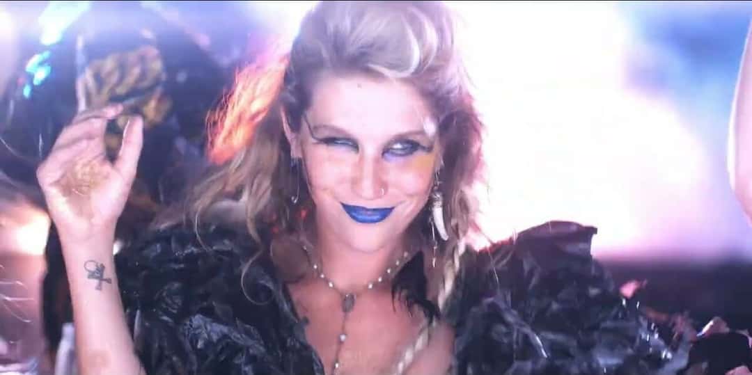Fotograma del video oficial de «Backstabber»: Kesha guiña un ojo, con labios azules, maquillaje dorado y un tapado de plumas negras, frente a luces rosas