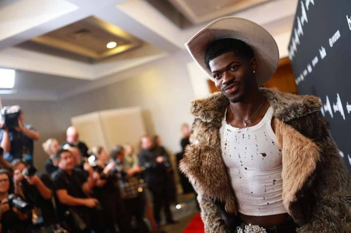 Lil Nas X sonriendo de perfil, con sombrero de cowboy claro, tapado de piel marrón y musculosa blanca gastada, frente a una fila de fotógrafos en una alfombra roja