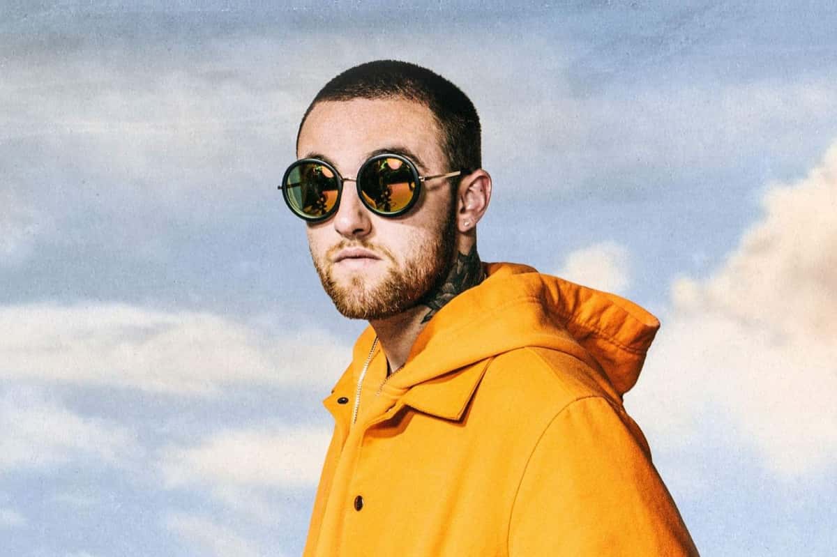 Mac Miller con campera naranja con capucha y anteojos de sol redondos espejados, delante de un fondo de cielo celeste con nubes