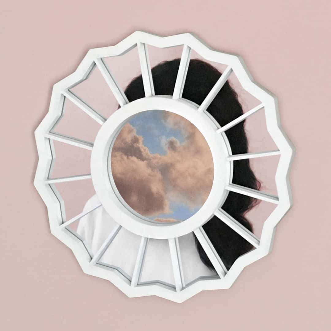 Portada original de The Divine Feminine de 2016: un marco blanco en forma de ventana redonda con un círculo de cielo y nubes en el centro, delante de una persona de pelo oscuro, sobre fondo rosa