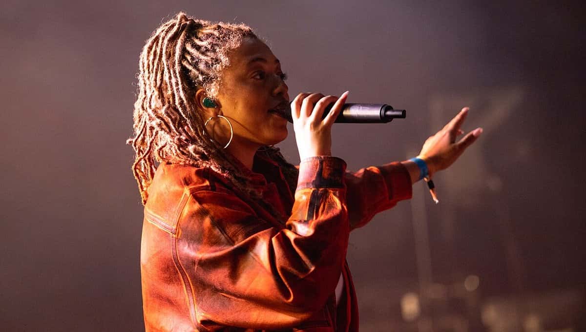 Mahalia canta con el micrófono en la mano derecha y el brazo izquierdo extendido, con rastas largas, aros grandes y campera marrón, en el festival Boardmasters (foto de archivo, agosto de 2021)