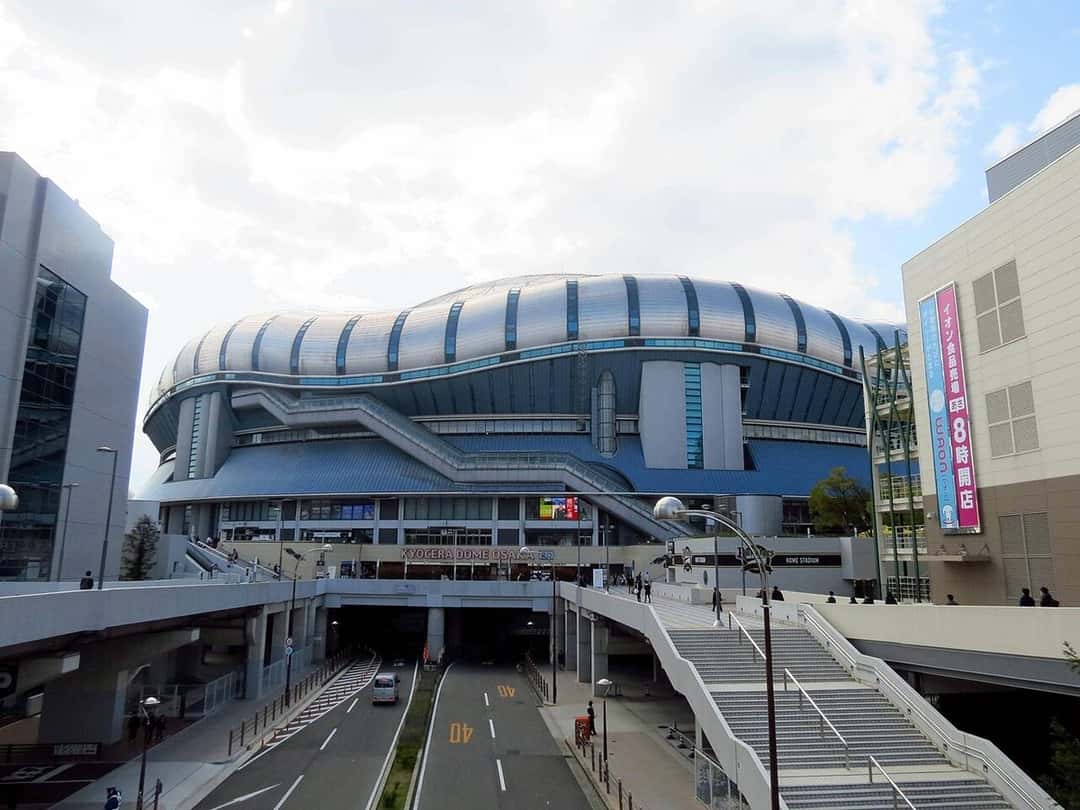 Vista del Kyocera Dome de Osaka de día, con su techo plateado en forma de domo, sobre una avenida con puentes peatonales y edificios a los costados (imagen del lugar de la ceremonia). Foto: redlegsfan21, CC BY-SA 2.0, vía Wikimedia Commons