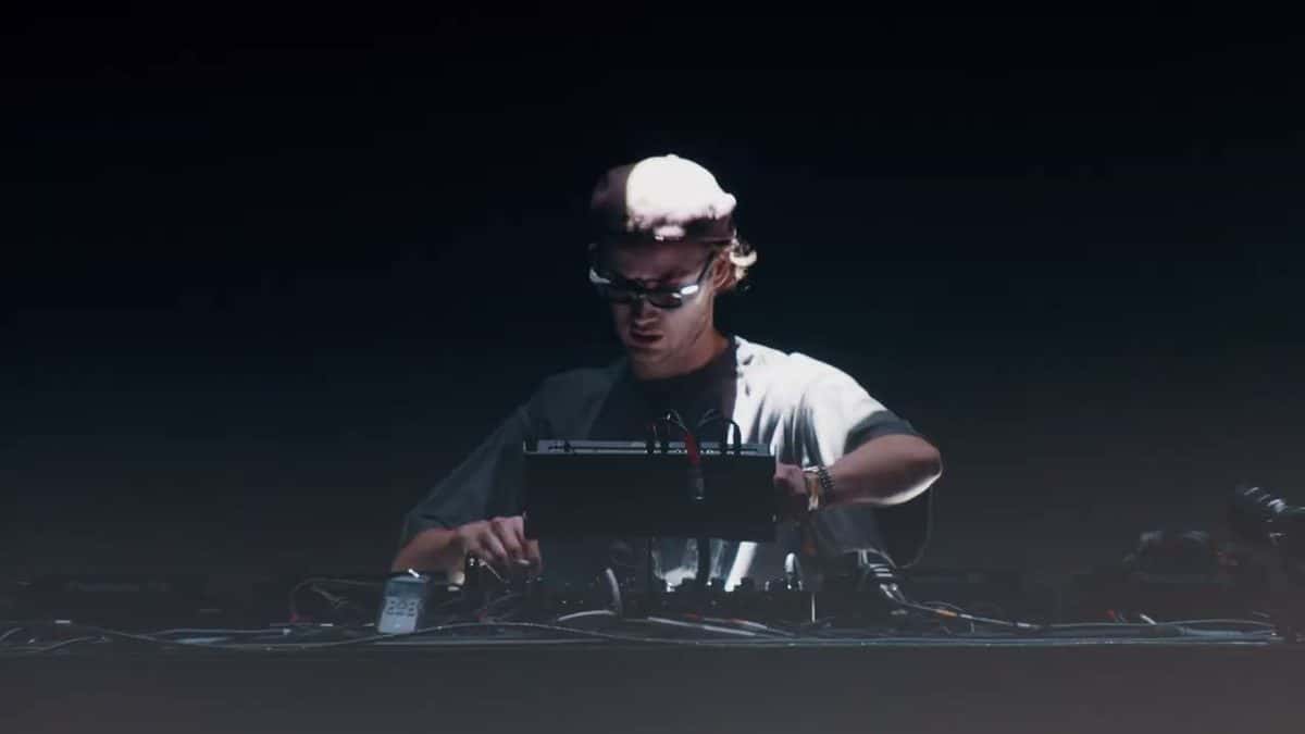 Fotograma del video oficial de Mau P en Coachella 2025: el DJ con gorra blanca y anteojos oscuros pincha detrás de la consola, sobre un fondo negro (foto de archivo)