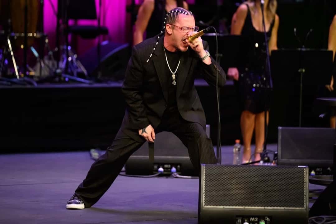 Mike Patton con traje negro y trenzas canta con las piernas abiertas y el micrófono dorado pegado a la boca, con las coristas de fondo, en el Palacio de Bellas Artes. Foto: Secretaría de Cultura