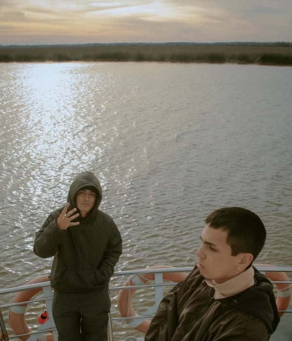 Milo J y Trueno sobre la cubierta de una embarcación, con el río y el atardecer de fondo, en una foto de prensa de La Jangada