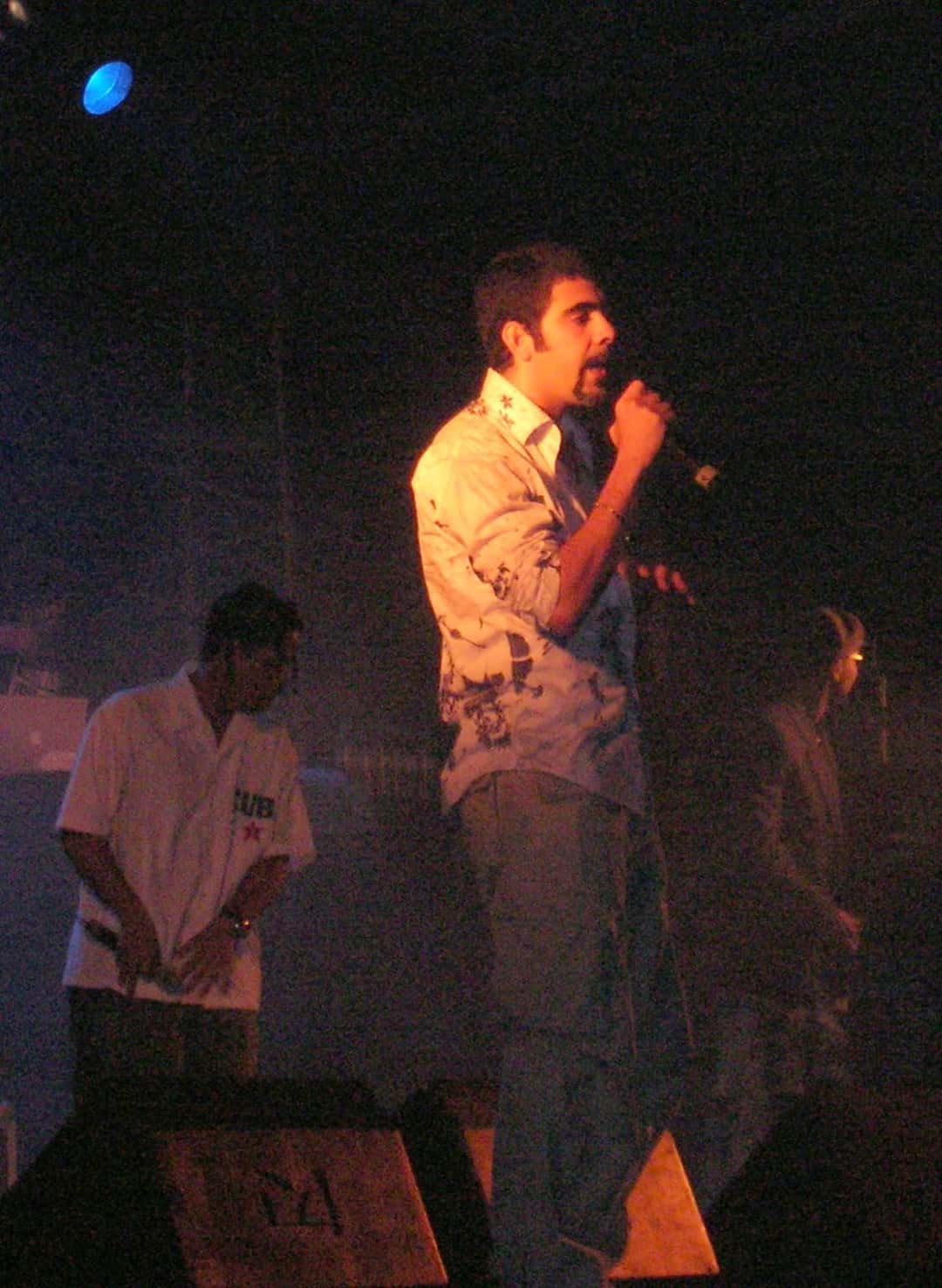 Orishas en vivo en Leioa, España, en junio de 2006: un integrante canta con micrófono bajo luces naranjas y otro lo acompaña en el escenario (foto de archivo). Foto: Javier Mediavilla Ezquibela, CC BY 2.5, vía Wikimedia Commons