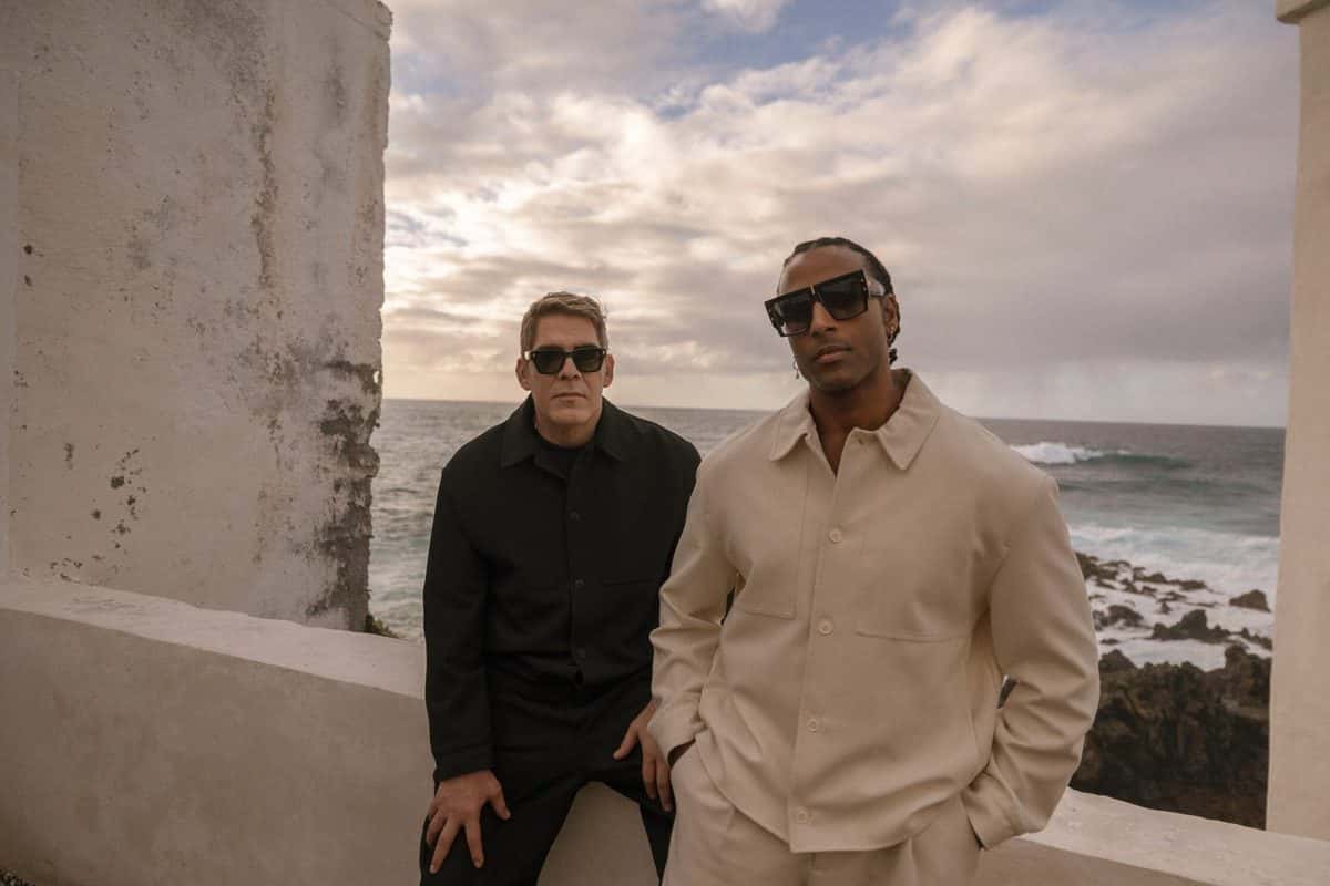 Foto de prensa de Orishas: Yotuel Romero y Roldán González, con anteojos de sol, sentados en un muro blanco frente al mar, uno de negro y el otro de blanco