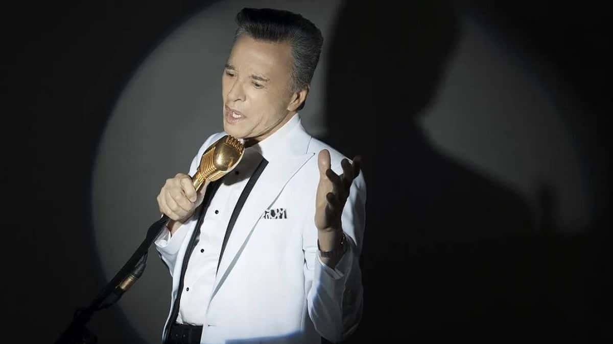 Palito Ortega canta con un micrófono dorado, de saco blanco y moño negro, iluminado por un círculo de luz sobre un fondo oscuro (foto de prensa de archivo)