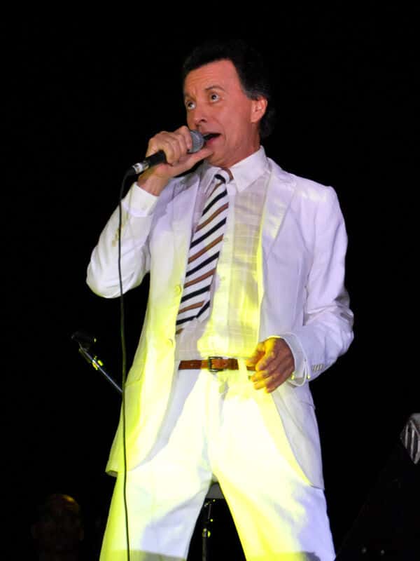 Palito Ortega canta con traje blanco y corbata a rayas, micrófono en mano, iluminado por una luz amarilla en un escenario oscuro, en un show de 2009 (foto de archivo). Foto: Damian Zanini, CC BY-SA 2.0, vía Wikimedia Commons