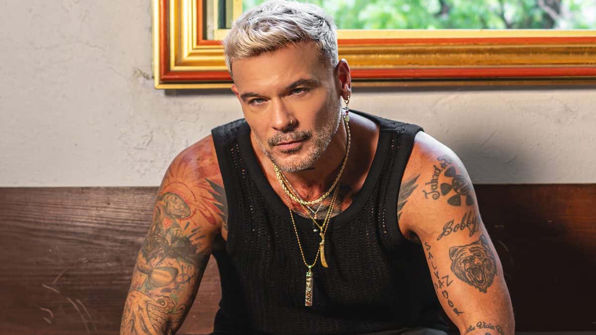 Foto de prensa de Pedro Capó: el cantante puertorriqueño, con pelo claro, barba, musculosa negra, collares y los brazos tatuados, sentado en un banco de madera frente a una pared blanca con un cuadro de marco dorado