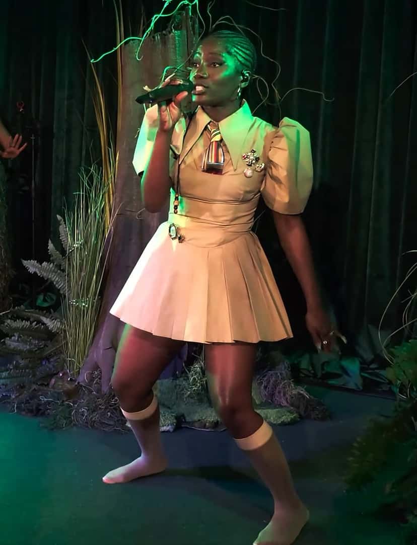 Foto de archivo de Doechii cantando en vivo con un micrófono en la mano, con vestido corto beige de mangas abullonadas y medias blancas, sobre un escenario con plantas y luces verdes. Foto: the CwE, Wikimedia Commons, CC BY 3.0