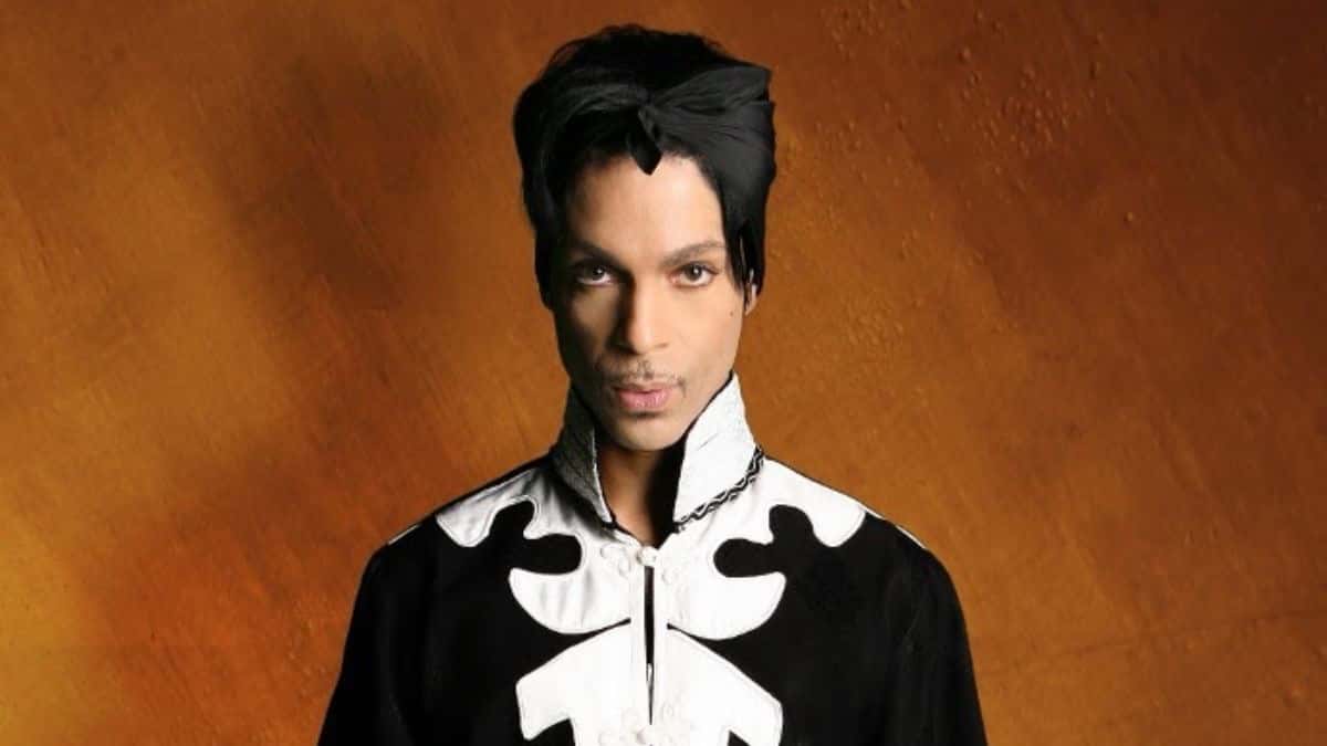 Foto de prensa de Prince en la época de «3121»: de frente, con pañuelo negro en la cabeza y una chaqueta negra de cuello alto con dibujos blancos, sobre un fondo anaranjado con textura
