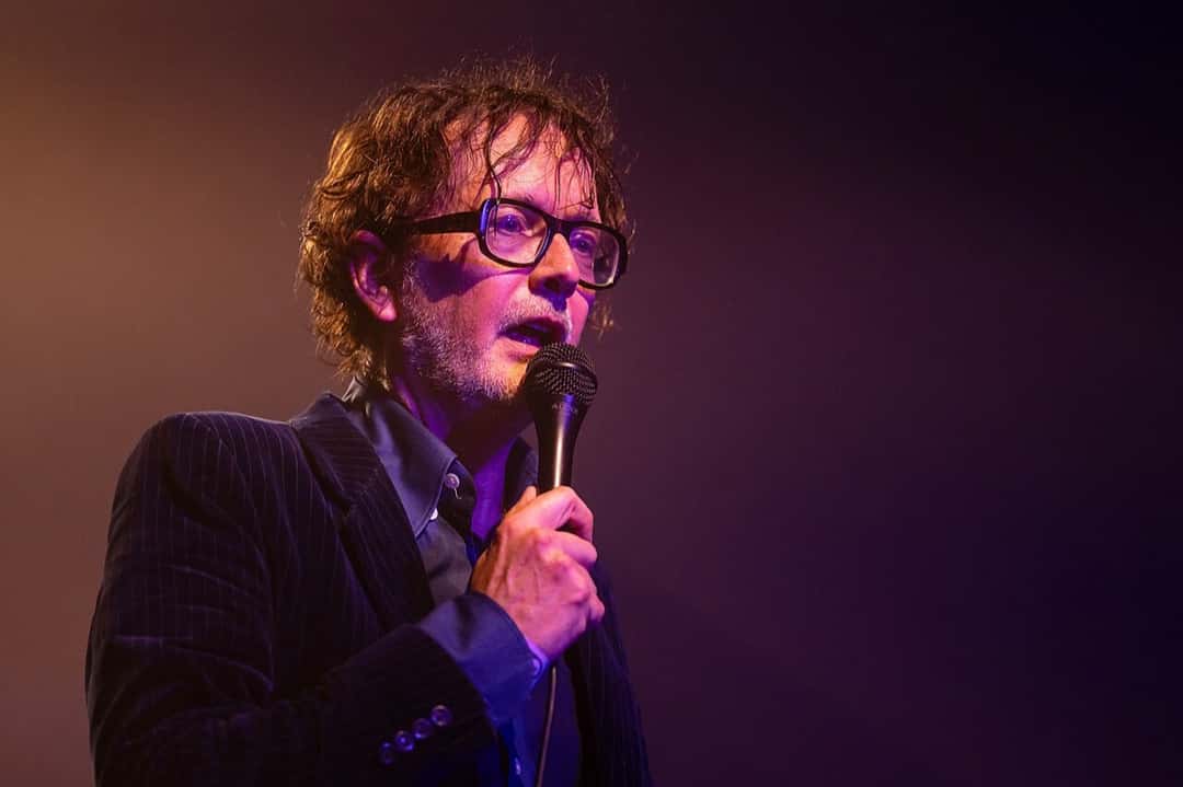 Jarvis Cocker canta con anteojos de marco grueso y saco a rayas, con el micrófono en la mano, bajo una luz violeta en el festival La Route du Rock de 2025 (foto de archivo). Foto: Selbymay, CC BY-SA 4.0, vía Wikimedia Commons