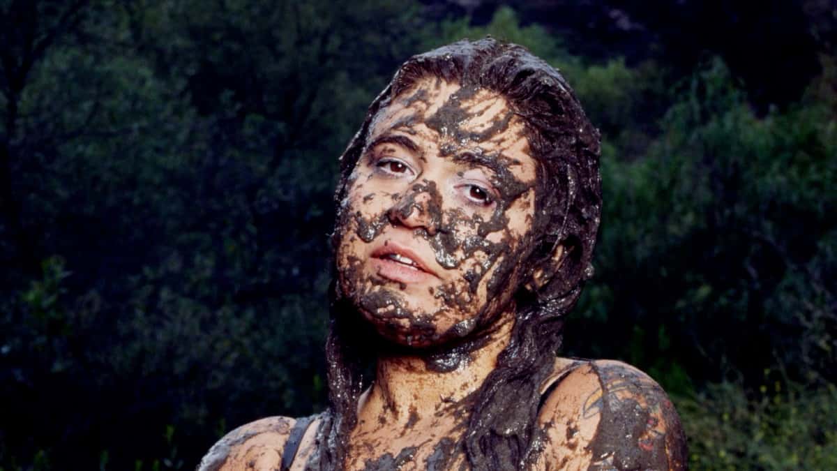 Foto de prensa oficial de Remi Wolf para "Mud": la cantante con la cara, el pelo y los hombros cubiertos de barro, frente a un fondo de árboles al atardecer