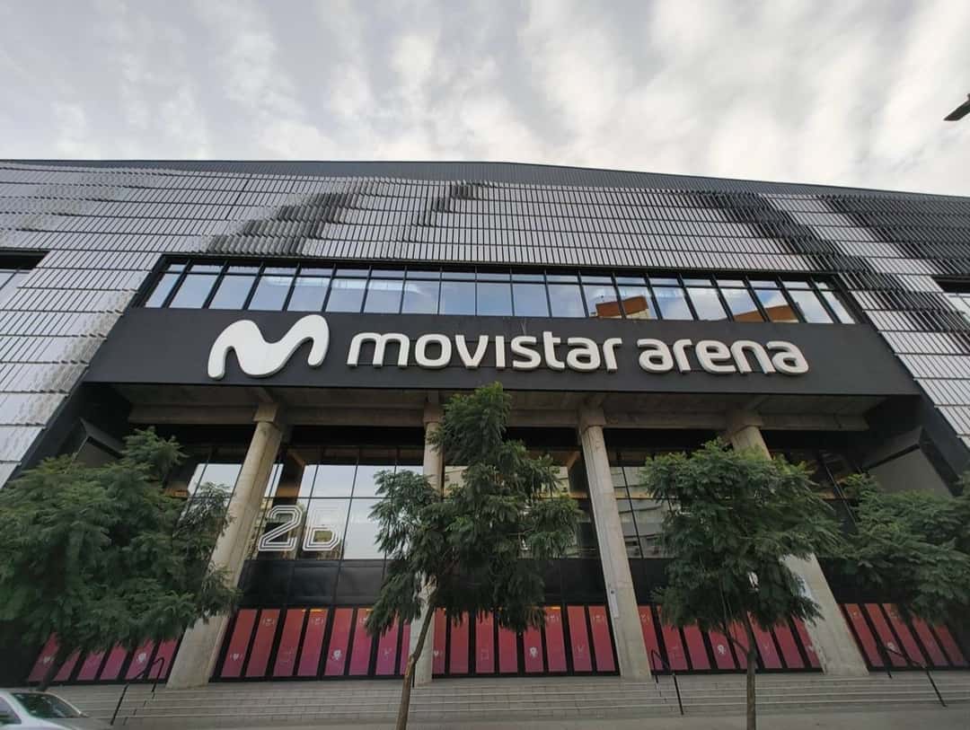 El frente del Movistar Arena de Buenos Aires, con el cartel del estadio sobre la entrada vidriada y árboles en la vereda (imagen del lugar del show). Foto: BugWarp, CC BY-SA 4.0, vía Wikimedia Commons
