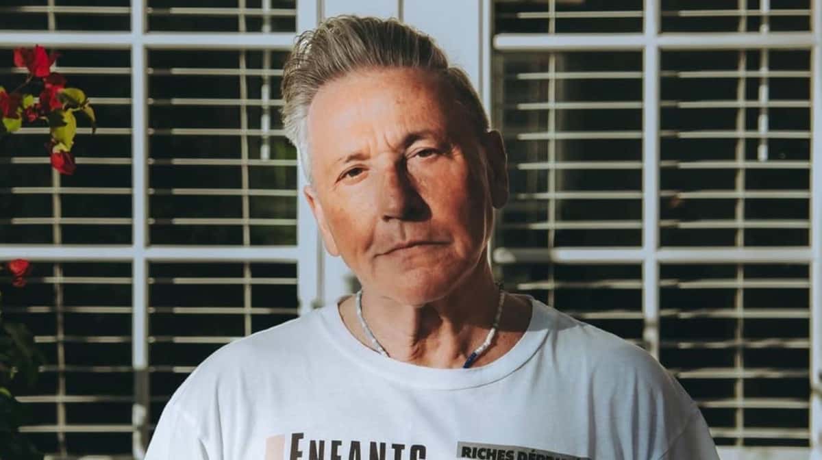 Ricardo Montaner, con pelo gris peinado hacia atrás, remera blanca y collares, mira a cámara de frente a una ventana con rejas blancas y flores rojas a la izquierda. Foto: Instagram de Ricardo Montaner