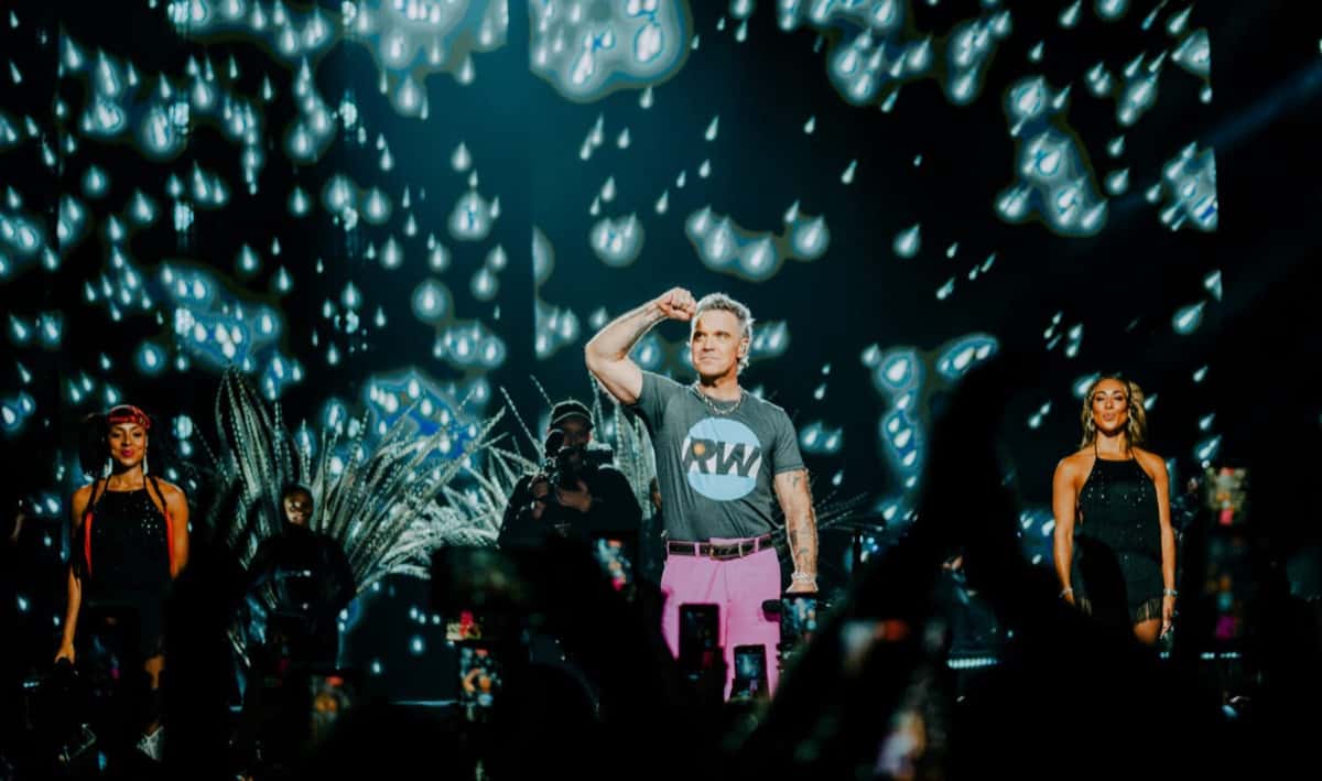 Robbie Williams en el escenario con remera gris con el logo RW y pantalón rosa, el puño cerrado a la altura de la frente, entre dos bailarinas vestidas de negro y frente a una pantalla con luces celestes