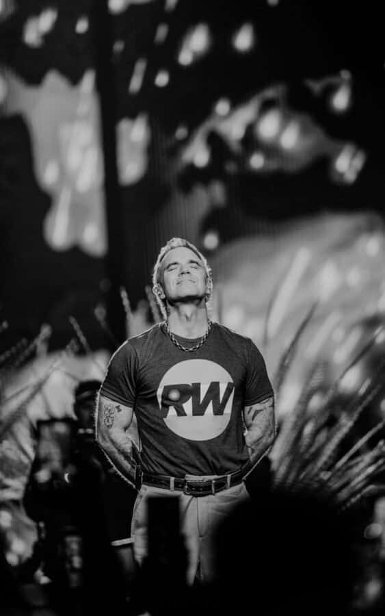 Robbie Williams en blanco y negro, de pie en el escenario con los ojos cerrados y una leve sonrisa, con remera con el logo RW, cadena al cuello y las manos atrás