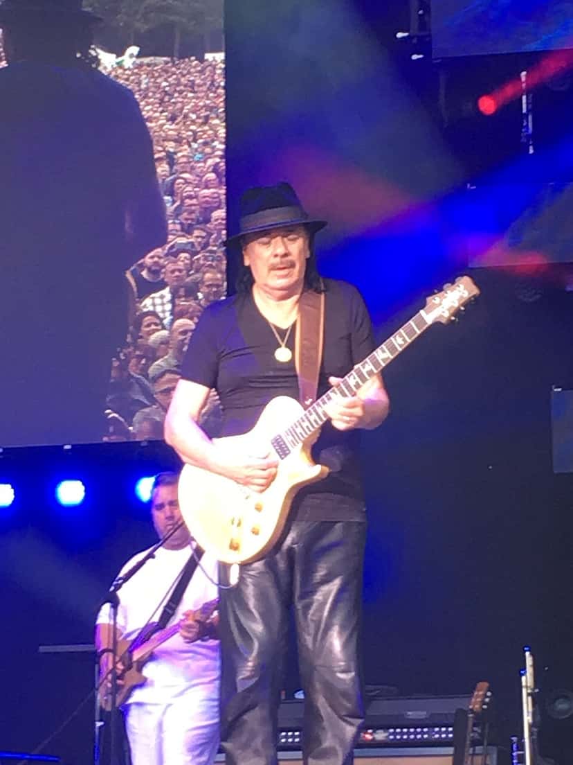 Carlos Santana toca una guitarra eléctrica clara con sombrero negro, remera negra y pantalón de cuero, con un músico de su banda detrás, en un show en Mainz, Alemania (foto de archivo, junio de 2018). Foto: Joho345, CC BY-SA 4.0, vía Wikimedia Commons