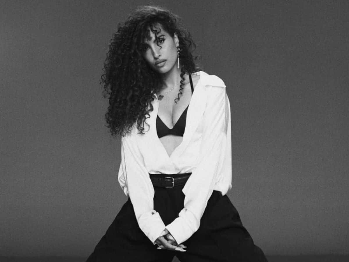 Foto de prensa de Snoh Aalegra en blanco y negro: la cantante, con pelo largo enrulado, camisa blanca abierta y pantalón oscuro, sentada sobre un fondo gris liso con las manos entrelazadas