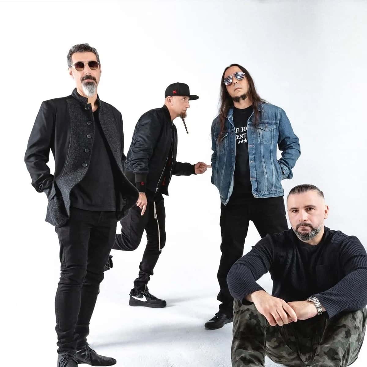 Foto de prensa de System of a Down: los cuatro integrantes, vestidos de negro y jean, posan sobre un fondo blanco, uno de ellos sentado en el piso