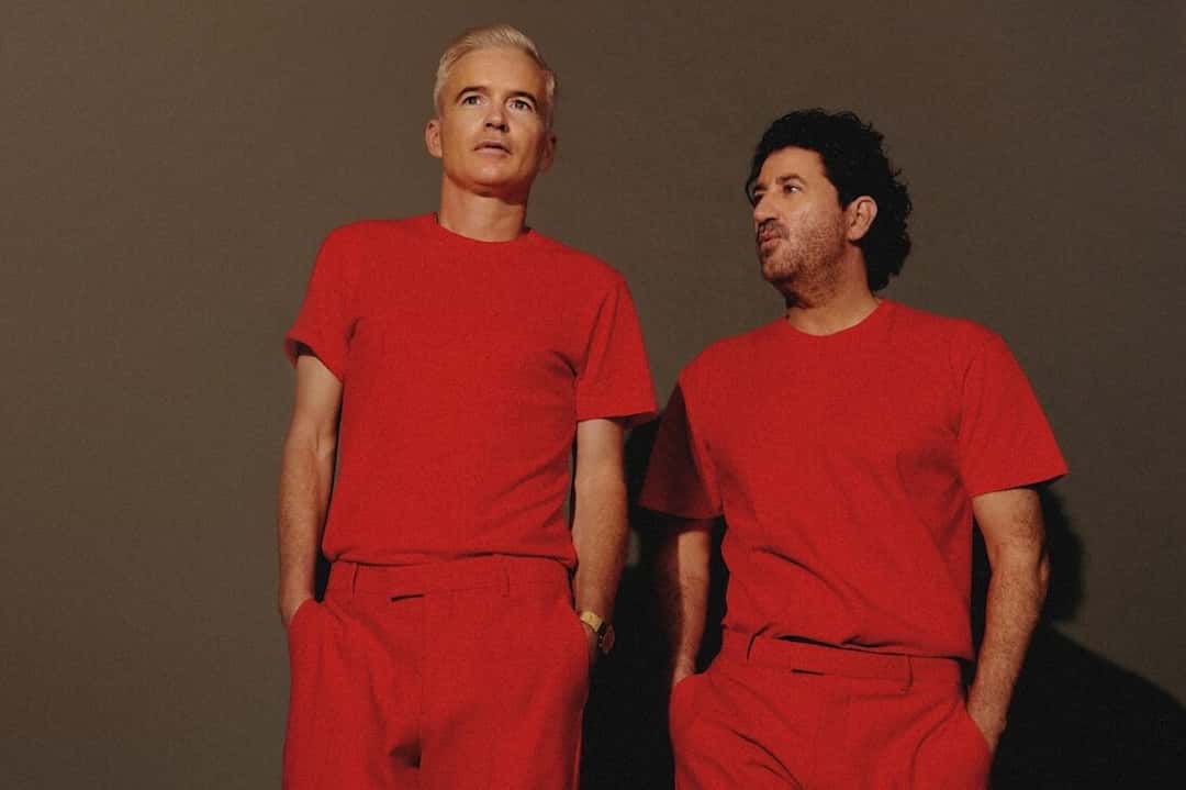 Dos integrantes de The Avalanches vestidos de rojo, con remera y pantalón, posando contra una pared marrón en una foto de prensa de la gira