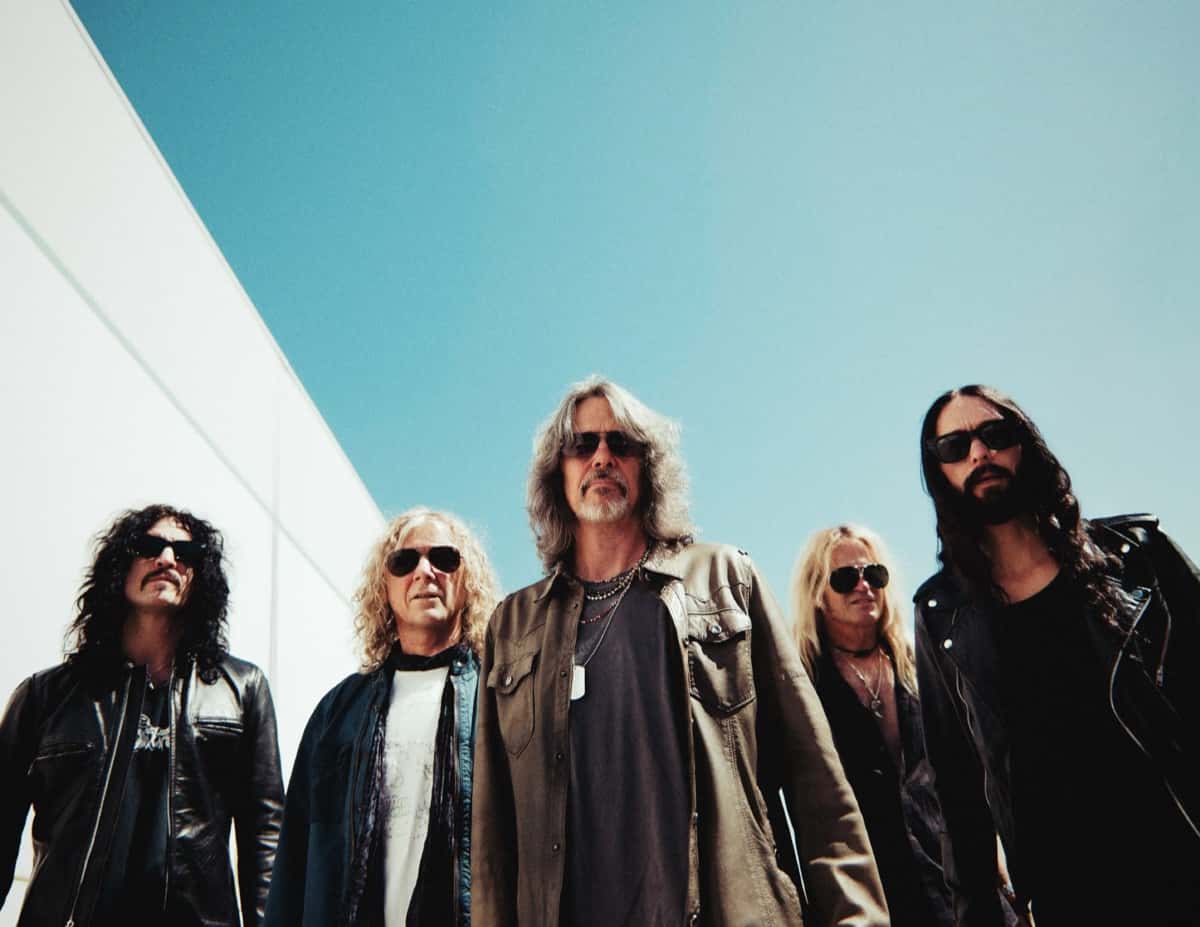 Foto de prensa de The Dead Daisies 2026: los cinco integrantes, con anteojos de sol y camperas de cuero y de gabardina, fotografiados desde abajo contra un cielo celeste y una pared blanca