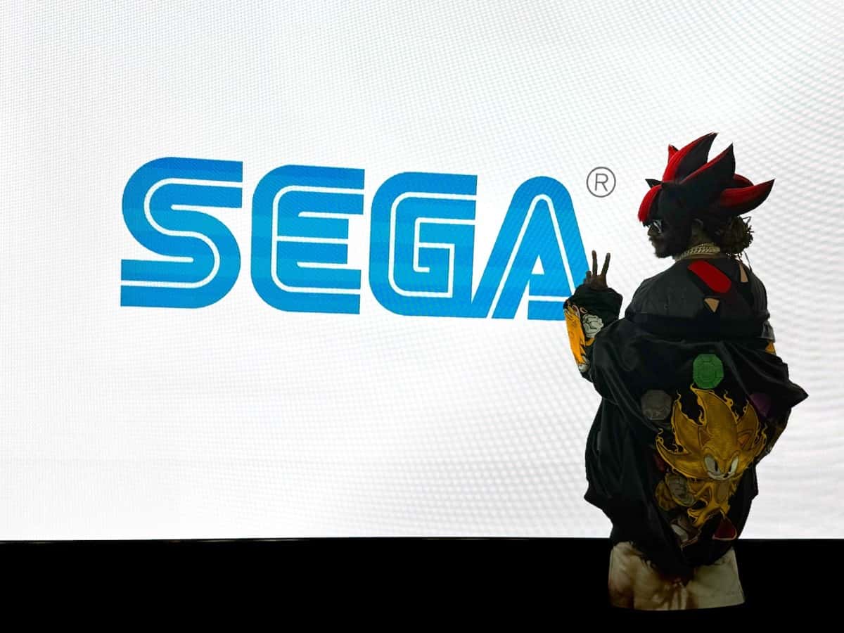 Foto oficial de Thundercat para "Moving At Light Speed": el músico, de espaldas y con un gorro con púas rojas y negras y una campera con Super Sonic bordado, hace la V con los dedos frente a una pantalla con el logo de SEGA