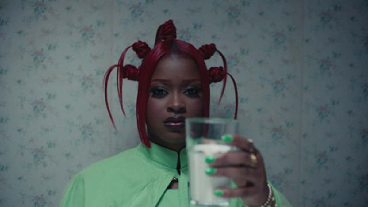 Imagen oficial del video de "CANDLE WAX" (foto de archivo, julio de 2026): Tierra Whack, con el pelo rojo atado en varios rodetes y mechones sueltos, campera verde clara y uñas verdes, mira a cámara y sostiene un vaso con un líquido blanco frente a una pared empapelada con flores