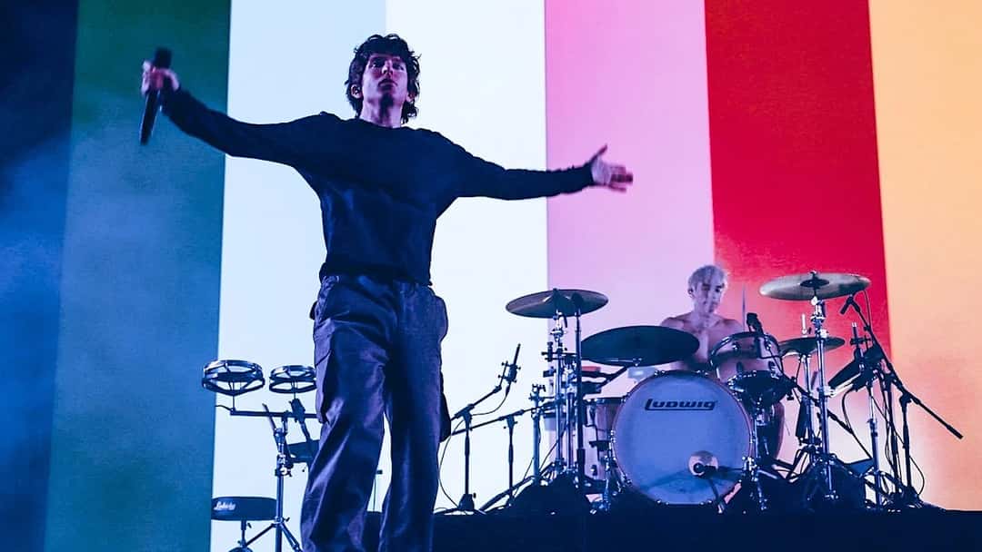El cantante de Turnstile, con buzo oscuro y micrófono en la mano, abre los brazos en el escenario; detrás, el baterista toca frente a un fondo de franjas de colores